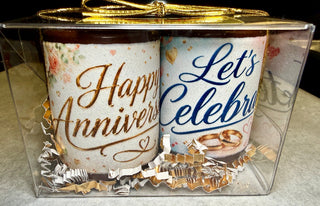 Set of 2 solid chocolate shot glasses with edible 'Happy Anniversary' in gold script and 'Let's Celebrate' in blue with floral and gold wedding ring prints, nestled in gold and white crinkle paper in a clear gift box with gold bow, Chamberlain's Chocolate, Atlanta GA