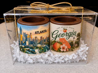 Set of 2 solid chocolate shot glasses in a clear gift box with gold bow — one with Atlanta skyline edible print, one with Georgia Peach State design, nestled in white crinkle paper from Chamberlains Chocolate in Atlanta