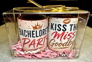 Set of 2 solid chocolate shot glasses with edible 'Bachelorette Party' with gold crown and 'Kiss the Miss Goodbye' with red lip print designs, nestled in pink and gold crinkle paper in a clear gift box with gold bow, Chamberlain's Chocolate, Atlanta GA