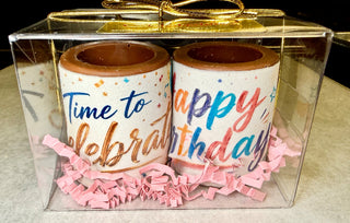 Two solid chocolate shot glasses with colorful multicolor edible 'Happy Birthday' and 'Time to Celebrate' prints, nestled in pink crinkle paper in a clear gift box with gold bow, made at Chamberlain's Chocolate, Atlanta GA