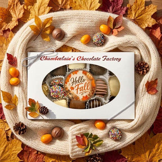 A boxed assortment of chocolates from Chamberlain's Chocolate Factory, wrapped in a white card with an oval window showing assorted truffles and chocolate pieces. The box is styled on a cream knit blanket surrounded by autumn leaves, pinecones, and small orange fruits.