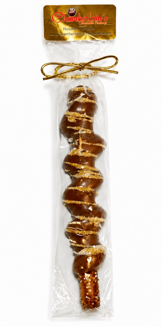 A packaged milk chocolate-covered pretzel rod with gold drizzle and sugar crystals, sealed in a clear plastic bag with a gold bow from Chamberlains Chocolate Factory