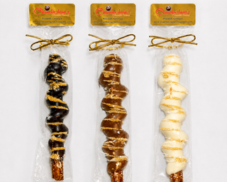 A packaged chocolate-covered pretzel rod with gold drizzle and sugar crystals, sealed in a clear plastic bag with a gold bow from Chamberlains Chocolate Factory