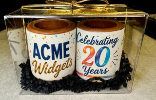 Top-angle view of 2 custom chocolate shot glasses with 'ACME Widgets' and 'Celebrating 20 Years' edible prints in black crinkle paper, clear acrylic gift box with gold bow, custom corporate event giveaway by Chamberlain's Chocolate, Atlanta GA