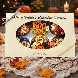 A boxed assortment of Halloween-themed chocolates from Chamberlain's Chocolate Factory, displayed on a table with autumn decorations, candles, pumpkins, and string lights in the background.