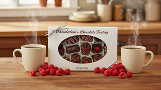 Chamberlain's Chocolate Factory raspberry chocolate and truffle gift box with oval window, displayed on a wooden table flanked by steaming coffee mugs and fresh raspberries, Atlanta Georgia