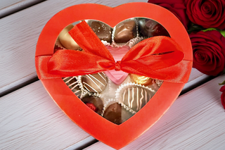 Heart-shaped box of chocolates with a red ribbon on a wooden surface with roses.

