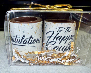 Set of 2 solid chocolate shot glasses with elegant black script edible 'Congratulations' and 'To The Happy Couple' prints, nestled in gold and white crinkle paper in a clear gift box with gold bow, made at Chamberlain's Chocolate, Atlanta GA