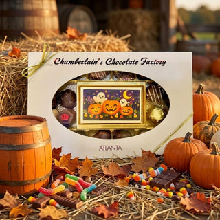 A boxed Halloween-themed chocolate gift set from Chamberlain's Chocolate Factory, displayed on straw with pumpkins, autumn leaves, candy corn, and barrels in the background. The white box has a clear oval window showing assorted chocolates and a central framed jack-o'-lantern illustration with ghosts and a crescent moon.
