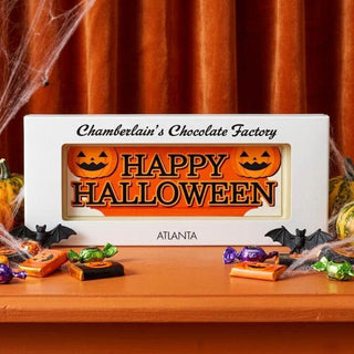 A personalized white chocolate gift box from Chamberlain's Chocolate Factory with 'HAPPY HALLOWEEN' printed in orange and black, styled on a wooden table with candy, bats, and cobwebs against an orange curtain backdrop.