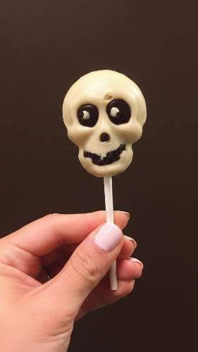 Hand holding a white chocolate skull-shaped lollipop with dark chocolate eyes and grinning mouth detail, artisan-made at Chamberlain's Chocolate in Roswell, Atlanta GA