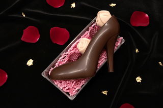 Dark chocolate high heel shoe filled with assorted truffles and white chocolate rose on dark velvet surface with rose petals and gold accents, luxury artisan chocolate gift flat lay