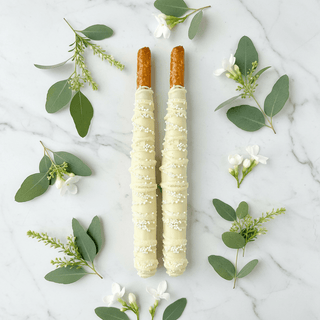 White chocolate-dipped pretzel rods with pearl sprinkles on marble surface with white flowers and greenery, premium artisan wedding confection flat lay