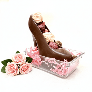 Artisan dark chocolate stiletto shoe with chocolate truffles and white chocolate rose in clear gift box with pink crinkle paper, styled with pink flowers, premium chocolate gift