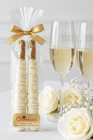 White chocolate-dipped pretzel rod wedding favors in clear bag with gold bow, displayed with champagne flutes and white roses, elegant wedding favor gift