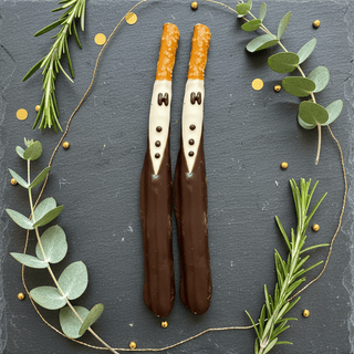 Two tuxedo-decorated dark chocolate pretzel rods with white chocolate bow tie and button details displayed on dark slate surface with greenery and gold accents, groom wedding favor flat lay