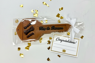Milk chocolate Key to Success gift in clear cellophane bag with white bow, flat lay on white surface with gold confetti and congratulations card, unique graduation or new job chocolate gift