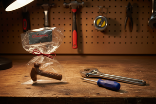 Hand-molded milk chocolate hammer in clear gift bag with red ribbon bow on wooden workbench next to real tools, unique Father's Day or handyman chocolate gift