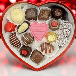 Heart-shaped box of assorted chocolates with a pink heart candy on a blurred red and white background.

